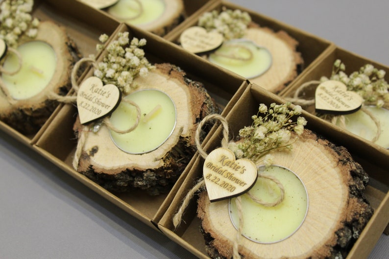 May include: A set of tea light candles in rustic wooden holders, arranged in a cardboard box. Each candle is surrounded by a wooden slice, dried flowers, and a heart-shaped tag with the text "Katie's Bridal Shower 8.22.2020".