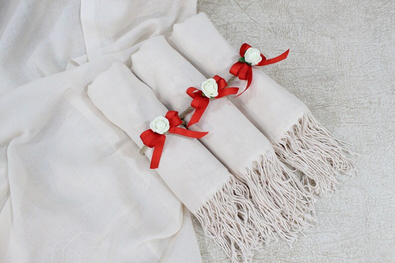 May include: Three white pashmina scarves with red ribbon bows and white flower accents. The scarves are folded and stacked on top of each other.