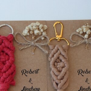 Puede incluir: Dos llaveros de macram&eacute; hechos a mano con un llavero de oro y plata. Los llaveros son rojos y marrones y tienen un patr&oacute;n de nudos decorativos. Los llaveros se muestran en una tarjeta marr&oacute;n con el texto "Robert & Lindsay" escrito en ella.