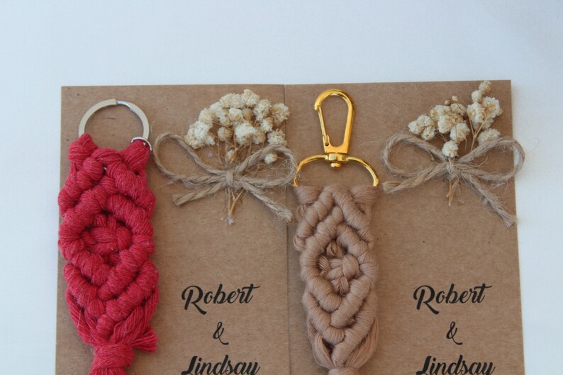 Puede incluir: Dos llaveros de macram&eacute; hechos a mano con un llavero de oro y plata. Los llaveros son rojos y marrones y tienen un patr&oacute;n de nudos decorativos. Los llaveros se muestran en una tarjeta marr&oacute;n con el texto "Robert & Lindsay" escrito en ella.