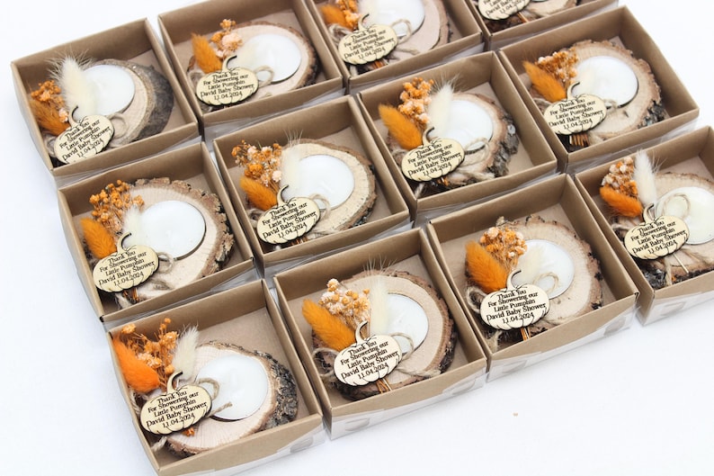 K&ouml;nnte beinhalten: Eine Sammlung von Babyparty-Gastgeschenken in kleinen Pappschachteln. Jede Schachtel enth&auml;lt eine Holzscheibe mit einer wei&szlig;en Kerze, Trockenblumen und einem Holzanh&auml;nger mit der Aufschrift "Thank You For Showering our Little Pumpkin David Baby Shower 12.04.2024."
