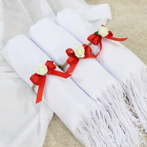 May include: Three white scarves with fringed edges are rolled up and tied with red ribbon bows and white flowers.