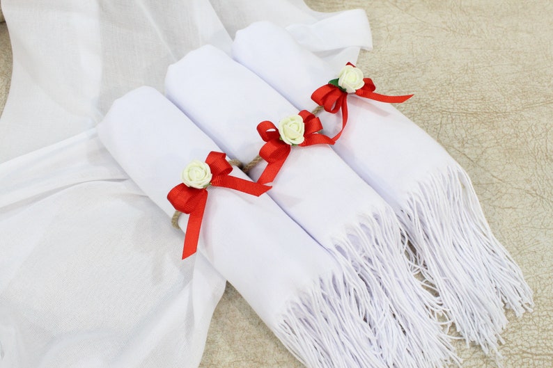 May include: Three white scarves with fringed edges are rolled up and tied with red ribbon bows and white flowers.