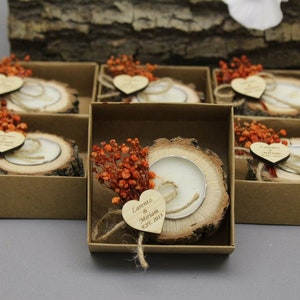 May include: A collection of rustic wedding favors. Each favor features a small candle set in a wooden slice, adorned with orange dried flowers, a wooden heart, and twine, presented in a small brown box.