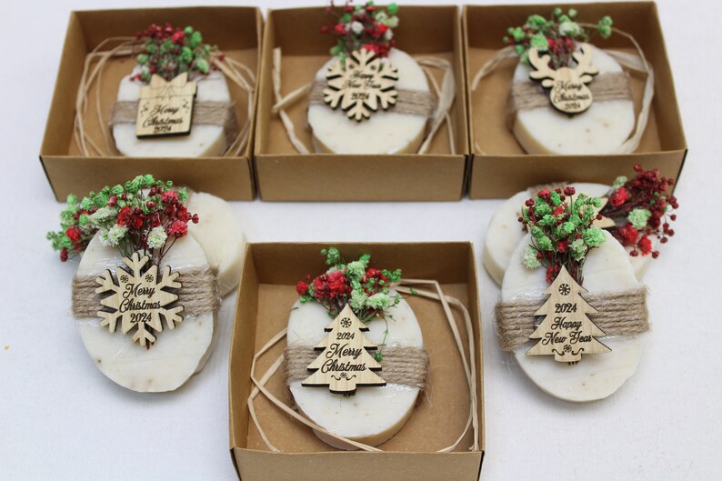 May include: Oval-shaped soaps with decorative wooden ornaments and dried flowers, presented in individual cardboard boxes. Each soap is wrapped with twine and features a different Christmas-themed wooden ornament with the text "Merry Christmas 2024" or "Happy New Year 2024".