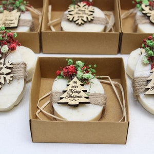 May include: Oval-shaped soaps, wrapped with twine and adorned with wooden Christmas ornaments and dried flowers, are presented in individual cardboard boxes. The ornaments feature the text "Merry Christmas 2024" and "Happy New Year 2024".