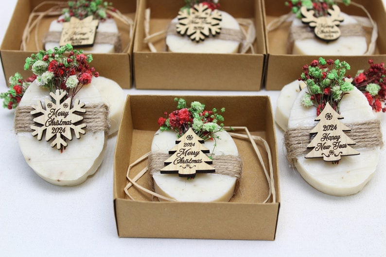 May include: Oval-shaped soaps, wrapped with twine and adorned with wooden Christmas ornaments and dried flowers, are presented in individual cardboard boxes. The ornaments feature the text "Merry Christmas 2024" and "Happy New Year 2024".