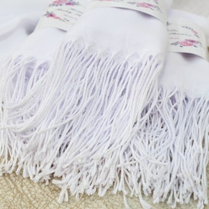May include: Two white pashmina scarves with long fringe. The scarves are folded and stacked on top of each other.