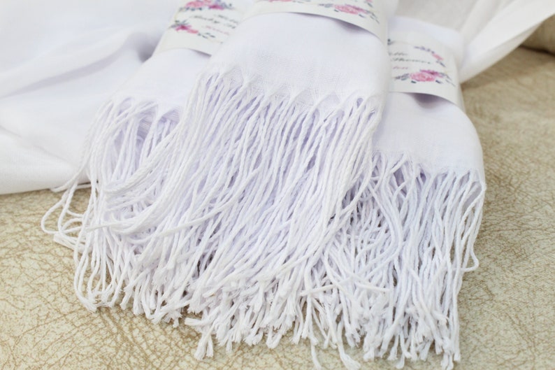 May include: Two white pashmina scarves with long fringe. The scarves are folded and stacked on top of each other.