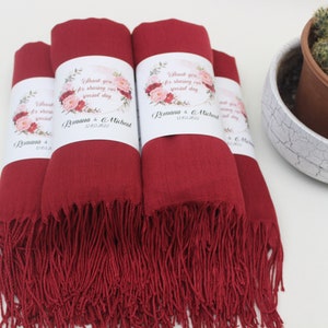 May include: Four burgundy red pashmina scarves with white floral wedding favour tags. The tags say "Thank you for sharing our special day" with the couple's names and wedding date.
