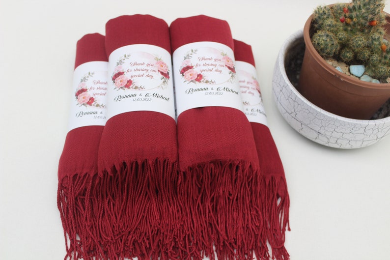 May include: Four burgundy red pashmina scarves with white floral wedding favour tags. The tags say "Thank you for sharing our special day" with the couple's names and wedding date.