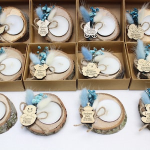 Op de afbeelding: Een set van 10 gepersonaliseerde babyshower-cadeautjes. Elk cadeautje is een klein houten schijfje met een teal blauwe kaars, gedroogde bloemen en een houten label met een babythema-ontwerp en gepersonaliseerde tekst. De labels bevatten een kinderwagen, een babyfles, een teddybeer, een baby-romper en een engel.