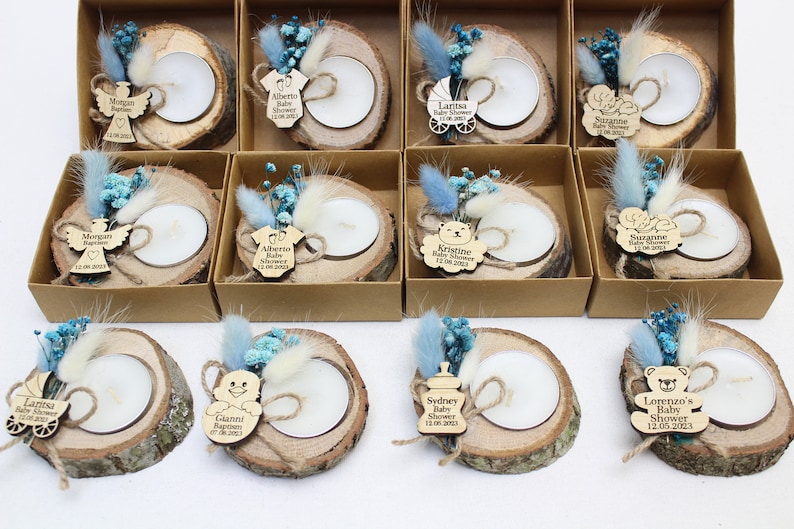 Op de afbeelding: Een set van 10 gepersonaliseerde babyshower-cadeautjes. Elk cadeautje is een klein houten schijfje met een teal blauwe kaars, gedroogde bloemen en een houten label met een babythema-ontwerp en gepersonaliseerde tekst. De labels bevatten een kinderwagen, een babyfles, een teddybeer, een baby-romper en een engel.