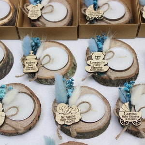 Op de afbeelding: Een verzameling kleine houten kaarshouders met theelichtjes, versierd met gedroogde bloemen, touw en gepersonaliseerde houten labels. De labels tonen babyshowerthema's met namen en data.