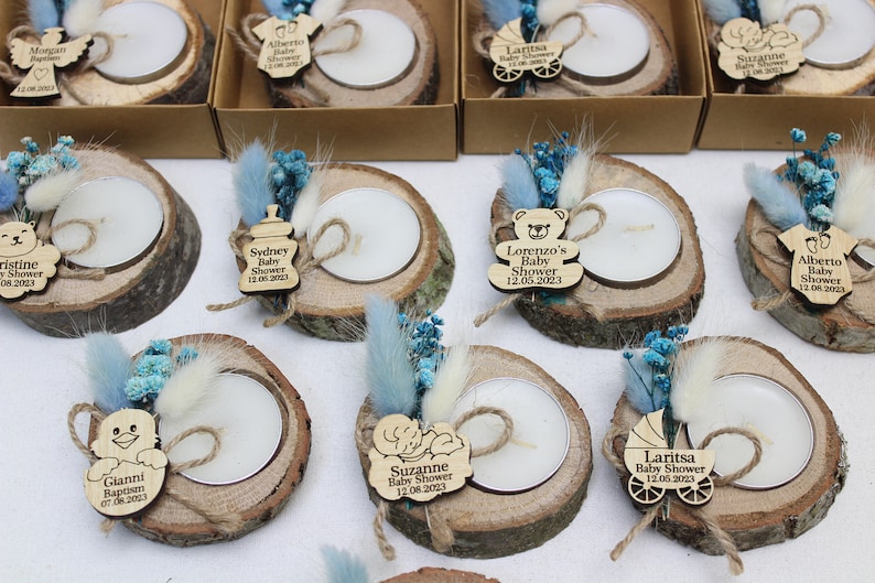 Op de afbeelding: Een verzameling kleine houten kaarshouders met theelichtjes, versierd met gedroogde bloemen, touw en gepersonaliseerde houten labels. De labels tonen babyshowerthema's met namen en data.