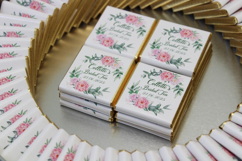 Puede incluir: Una bandeja de tabletas de chocolate envueltas individualmente con un dise&ntilde;o floral y el texto "Collette's Bridal Tea" y una fecha. Las tabletas de chocolate est&aacute;n envueltas en papel blanco con detalles de l&aacute;mina dorada.