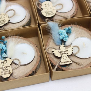 Op de afbeelding: Een set kleine, gepersonaliseerde houten kaarshouders met theelichtjes, blauwe en witte gedroogde bloemen en een houten label met een naam en datum. De labels tonen een baby-romper, een engel en een kinderwagen.
