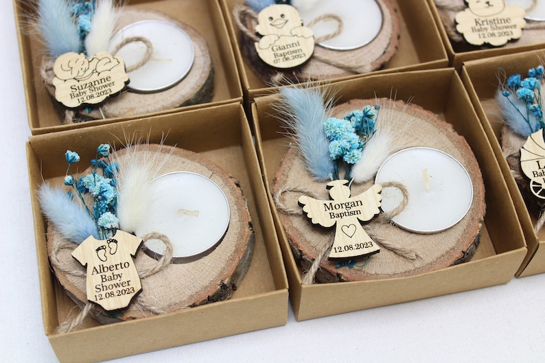 Op de afbeelding: Een set kleine, gepersonaliseerde houten kaarshouders met theelichtjes, blauwe en witte gedroogde bloemen en een houten label met een naam en datum. De labels tonen een baby-romper, een engel en een kinderwagen.