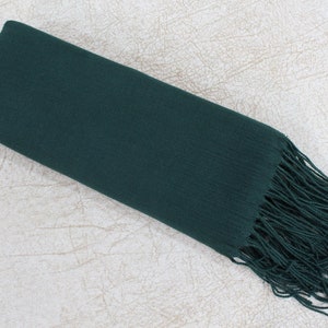 May include: A dark green scarf with long fringe on one end. The scarf is folded in half and lying on a white textured surface.
