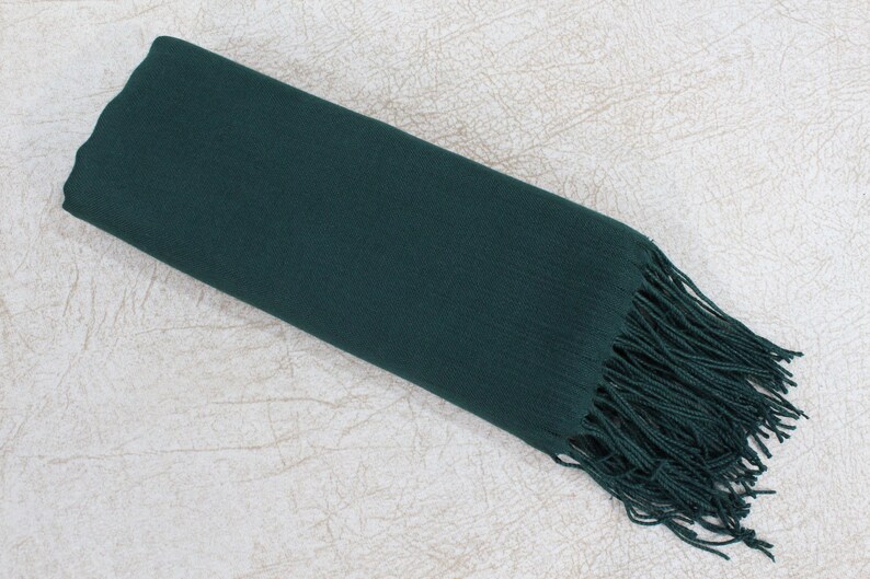 May include: A dark green scarf with long fringe on one end. The scarf is folded in half and lying on a white textured surface.