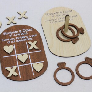 K&ouml;nnte beinhalten: Holz-Hochzeitsspiel-Set, einschlie&szlig;lich eines Tic-Tac-Toe-Bretts mit Herzen und Kreuzen, einem Ringwurfspiel und zwei ringf&ouml;rmigen Dekorationen. Das Set ist mit einem Hochzeitsdatum und einer Dankesbotschaft graviert.