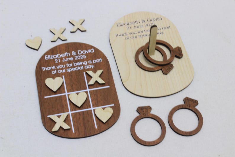 K&ouml;nnte beinhalten: Holz-Hochzeitsspiel-Set, einschlie&szlig;lich eines Tic-Tac-Toe-Bretts mit Herzen und Kreuzen, einem Ringwurfspiel und zwei ringf&ouml;rmigen Dekorationen. Das Set ist mit einem Hochzeitsdatum und einer Dankesbotschaft graviert.