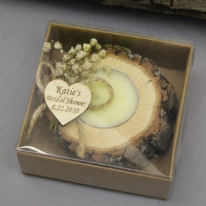 Op de afbeelding: Een klein houten doosje met een doorzichtig deksel, dat een houten kaarsenhouder met een kleine witte kaars bevat. De kaarsenhouder is versierd met gedroogde bloemen en een hartvormig houten label met de tekst "Katie's Bridal Shower 8.22.2020".
