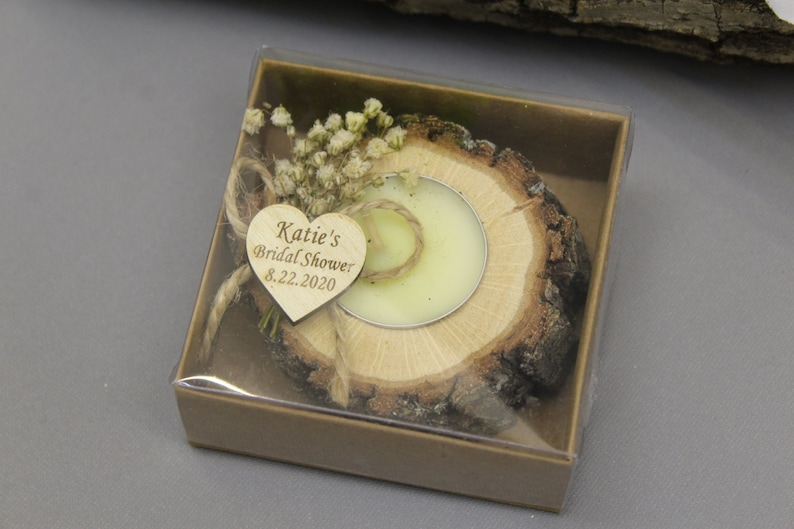 Op de afbeelding: Een klein houten doosje met een doorzichtig deksel, dat een houten kaarsenhouder met een kleine witte kaars bevat. De kaarsenhouder is versierd met gedroogde bloemen en een hartvormig houten label met de tekst "Katie's Bridal Shower 8.22.2020".