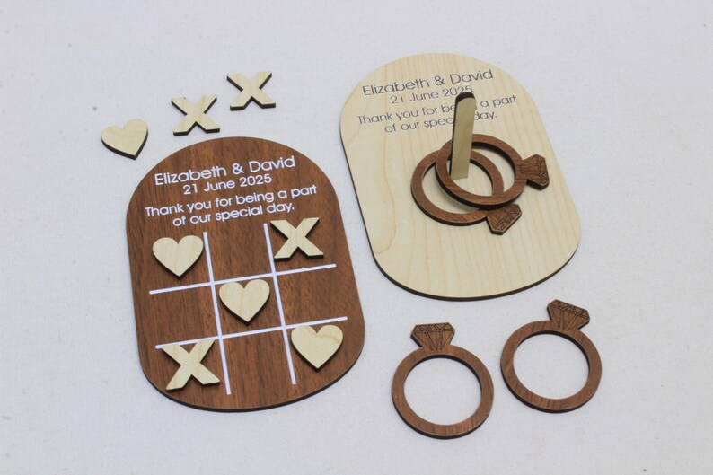 K&ouml;nnte beinhalten: Holz-Hochzeitsspiel-Set, einschlie&szlig;lich eines Tic-Tac-Toe-Bretts mit Herzen und X'en, einem Ringwurfspiel und zwei ringf&ouml;rmigen Dekorationen. Das Set tr&auml;gt die Namen "Elizabeth & David" und das Datum "21. Juni 2025".