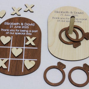 May include: Wooden wedding favors featuring a tic-tac-toe game board and a ring toss game. The tic-tac-toe board is dark brown with white lines and includes hearts and X's. The ring toss game has interlocking rings. Text on the favors reads "Elizabeth & David 21 June 2025".