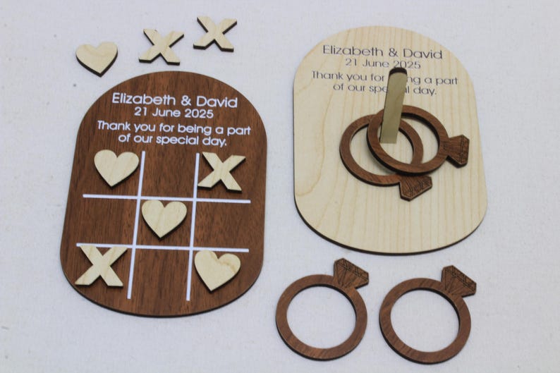 May include: Wooden wedding favors featuring a tic-tac-toe game board and a ring toss game. The tic-tac-toe board is dark brown with white lines and includes hearts and X's. The ring toss game has interlocking rings. Text on the favors reads "Elizabeth & David 21 June 2025".