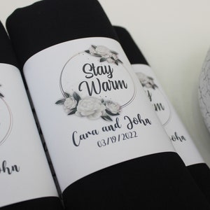 May include: Two black fabric gift bags with white labels. The labels have a floral design and the text "Stay Warm" with a gold circle around the text. The labels also include the names "Cara and John" and the date "03/19/2022".