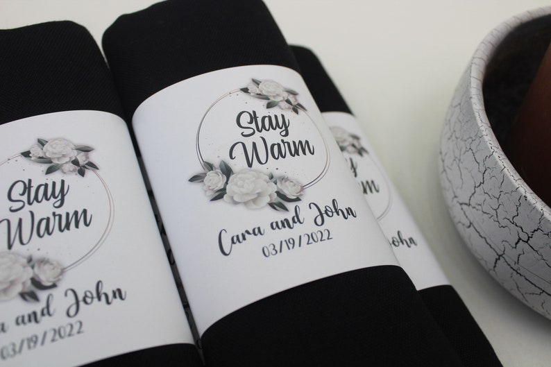 May include: Two black fabric gift bags with white labels. The labels have a floral design and the text "Stay Warm" with a gold circle around the text. The labels also include the names "Cara and John" and the date "03/19/2022".