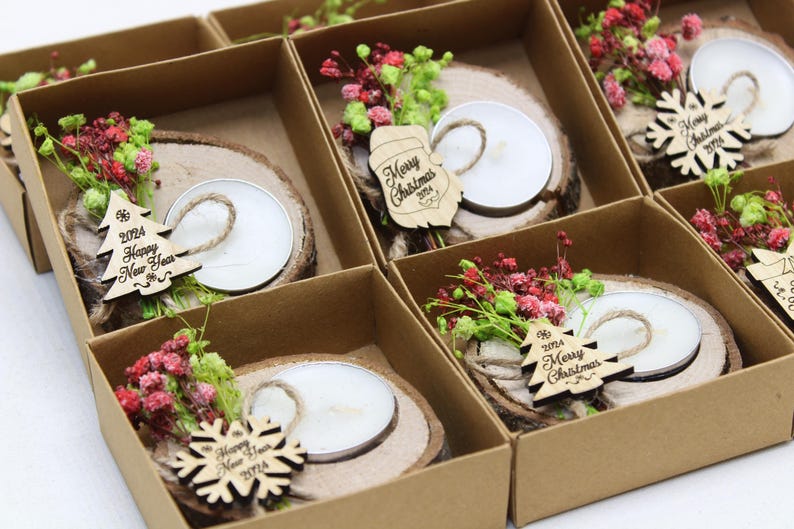 May include: An assortment of small, brown cardboard boxes with lids, each containing a wooden slice with a small white candle, a sprig of dried flowers, and a wooden ornament with a Christmas or New Year's message. The ornaments include a snowflake, a Christmas tree, and a New Year's tree.