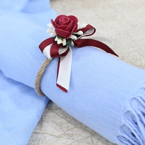 Pu&ograve; includere: Una sciarpa azzurra con un nastro bordeaux e bianco legato intorno. Una rosa bordeaux con dettagli bianchi &egrave; attaccata al nastro.
