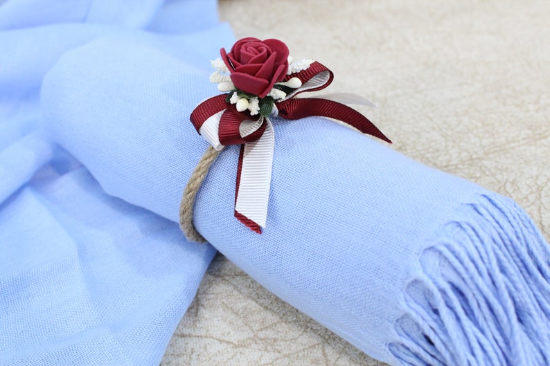 Pu&ograve; includere: Una sciarpa azzurra con un nastro bordeaux e bianco legato intorno. Una rosa bordeaux con dettagli bianchi &egrave; attaccata al nastro.