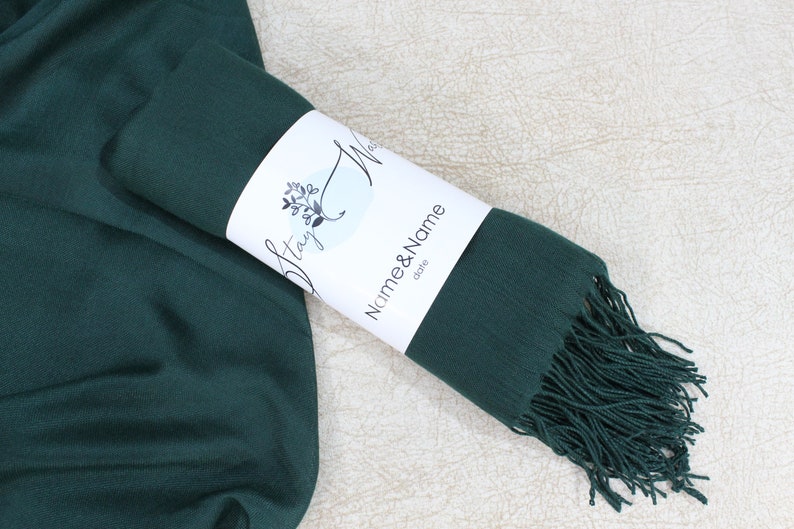 May include: A dark green scarf with fringe on the end. The scarf is rolled up and tied with a white label that says "Stay Wild" and "Name & Name" with the word "gifts" underneath.
