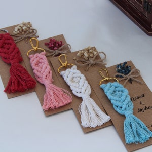 Puede incluir: Cuatro llaveros de macram&eacute; hechos a mano con borlas en rojo, rosa, blanco y azul. Cada llavero est&aacute; unido a un anillo de llave dorado y se muestra en una tarjeta marr&oacute;n con una cinta blanca y flores secas. La tarjeta del llavero azul tiene el texto "Robert & Lindsay 10.23.2020 &iexcl;Gracias por celebrar con nosotros!"