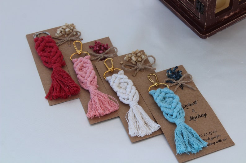 Puede incluir: Cuatro llaveros de macram&eacute; hechos a mano con borlas en rojo, rosa, blanco y azul. Cada llavero est&aacute; unido a un anillo de llave dorado y se muestra en una tarjeta marr&oacute;n con una cinta blanca y flores secas. La tarjeta del llavero azul tiene el texto "Robert & Lindsay 10.23.2020 &iexcl;Gracias por celebrar con nosotros!"