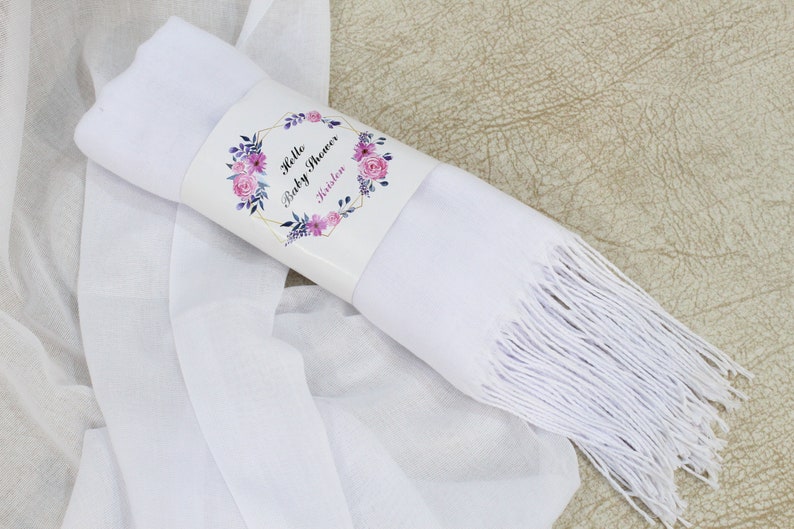 May include: White pashmina scarf with a fringe border. The scarf is rolled up and tied with a label that says "Hello Baby Shower" with a floral design.