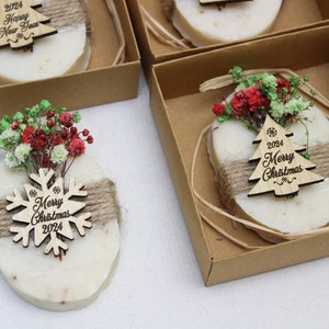 May include: Oval-shaped soaps with a natural color, decorated with a wooden Christmas tree or snowflake ornament. Each soap is wrapped with twine and topped with dried flowers, presented in a small brown box.