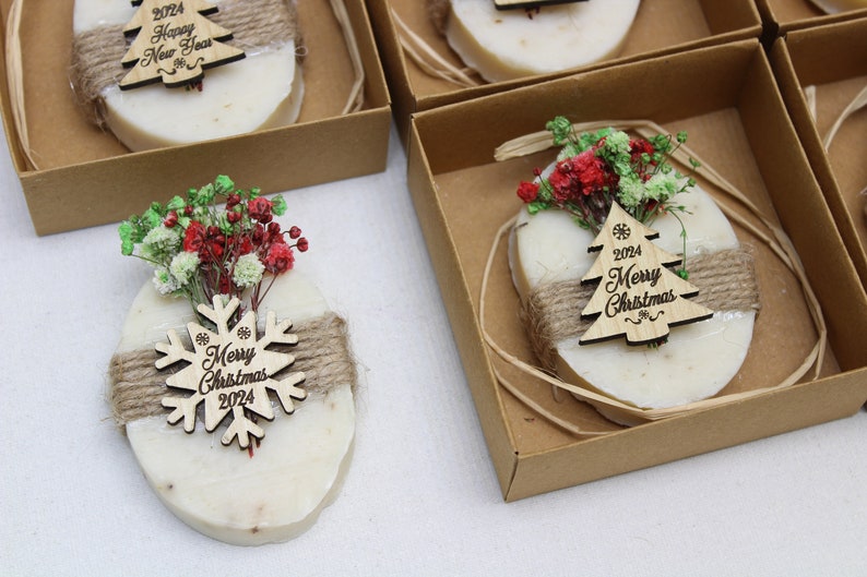 May include: Oval-shaped soaps with a natural color, decorated with a wooden Christmas tree or snowflake ornament. Each soap is wrapped with twine and topped with dried flowers, presented in a small brown box.