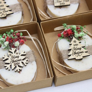 May include: Oval-shaped soaps with twine and wooden Christmas tree or snowflake ornaments, packaged in brown boxes. Each soap is decorated with dried flowers and the text "Merry Christmas 2024" or "Happy New Year 2024".
