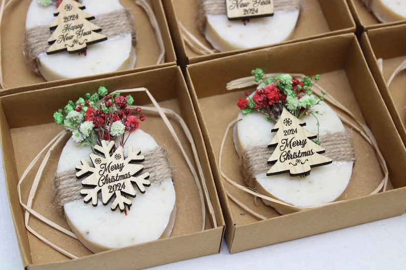 May include: Oval-shaped soaps with twine and wooden Christmas tree or snowflake ornaments, packaged in brown boxes. Each soap is decorated with dried flowers and the text "Merry Christmas 2024" or "Happy New Year 2024".