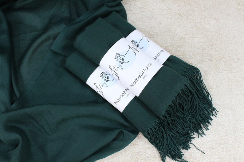 May include: Three dark green pashmina scarves with fringed edges, stacked and tied with a white label that reads "Name & Name" and "Stay"