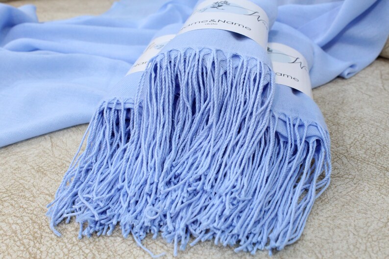 Pu&ograve; includere: Una sciarpa di pashmina blu chiaro con frange. La sciarpa &egrave; piegata e ha un'etichetta attaccata che dice "Name & Name".