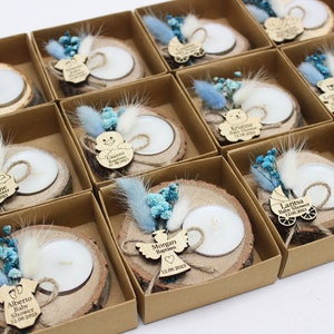 Op de afbeelding: Een verzameling kleine, rustieke houten kaarsenhouders met gepersonaliseerde labels. Elke houder heeft een kleine, witte kaars, een houten uitsnijding en gedroogde blauwe en witte bloemen. De labels bevatten namen en data, zoals "Morgan Doop 12.08.2023" en "Laritsa Baby Shower 12.08.2023".