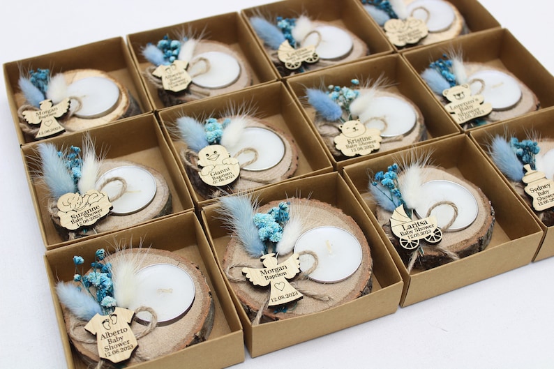 Op de afbeelding: Een verzameling kleine, rustieke houten kaarsenhouders met gepersonaliseerde labels. Elke houder heeft een kleine, witte kaars, een houten uitsnijding en gedroogde blauwe en witte bloemen. De labels bevatten namen en data, zoals "Morgan Doop 12.08.2023" en "Laritsa Baby Shower 12.08.2023".