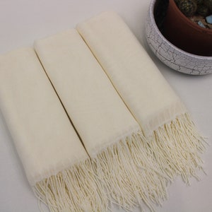 May include: Three ivory white scarves with fringe edges. The scarves are folded and stacked on top of each other.