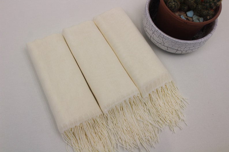 May include: Three ivory white scarves with fringe edges. The scarves are folded and stacked on top of each other.